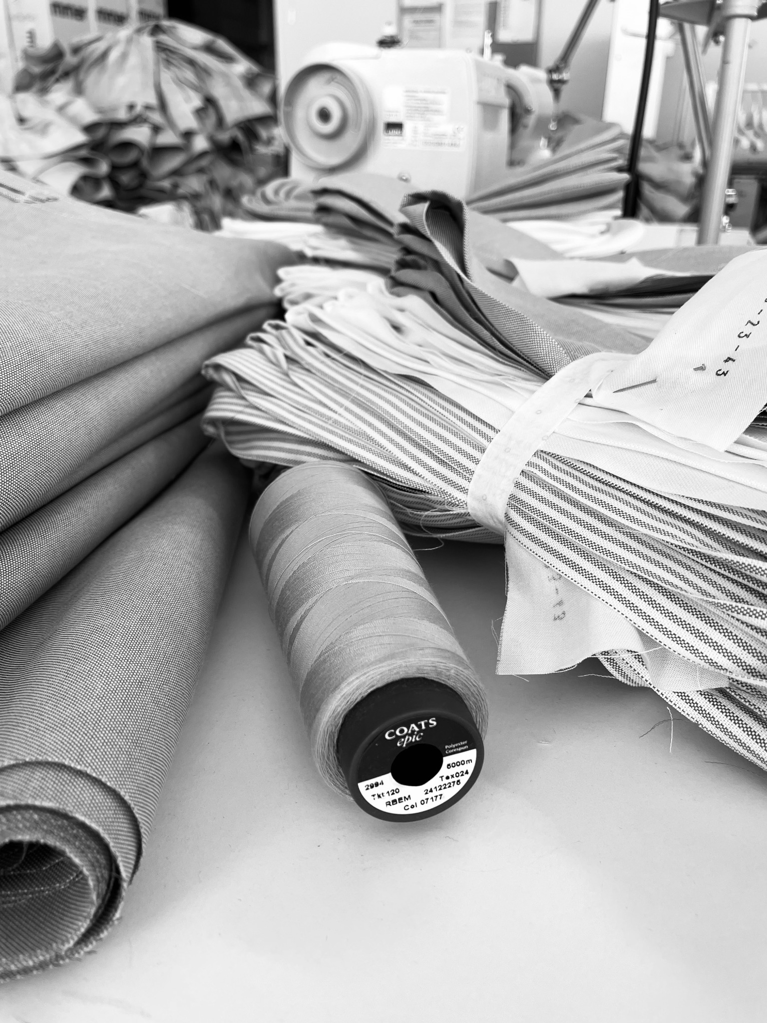 Czarno-białe ujęcie stosu tkanin w rolkach i pociętych pasków, szpulki nici Coats Epic oraz maszyny do szycia w tle.