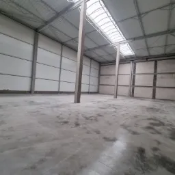 Wnętrze hali produkcyjnej z betonową posadzką, widoczne słupy konstrukcyjne i świetliki w dachu, ściany z paneli warstwowych.