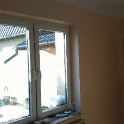 Pokój przed malowaniem: beżowe ściany, białe okno z widokiem na podwórze i ciemne drzwi z szybkami.