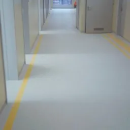 Długi korytarz w budynku użyteczności publicznej z szarą posadzką i żółtymi liniami przy ścianach, widoczne drzwi i okno na końcu korytarza, oświetlenie sufitowe.