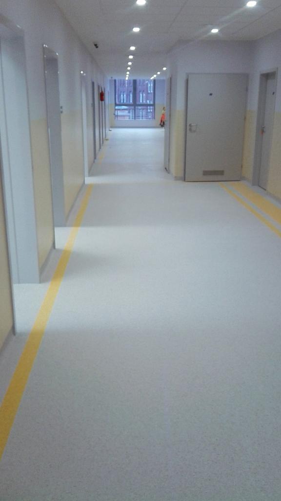 Długi korytarz w budynku użyteczności publicznej z szarą posadzką i żółtymi liniami przy ścianach, widoczne drzwi i okno na końcu korytarza, oświetlenie sufitowe.