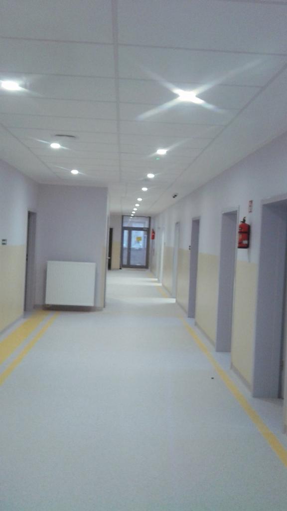 Długi, jasny korytarz z rzędem punktowych świateł w suficie, żółtymi pasami na ścianach i podłodze, oraz grzejnikiem na ścianie.