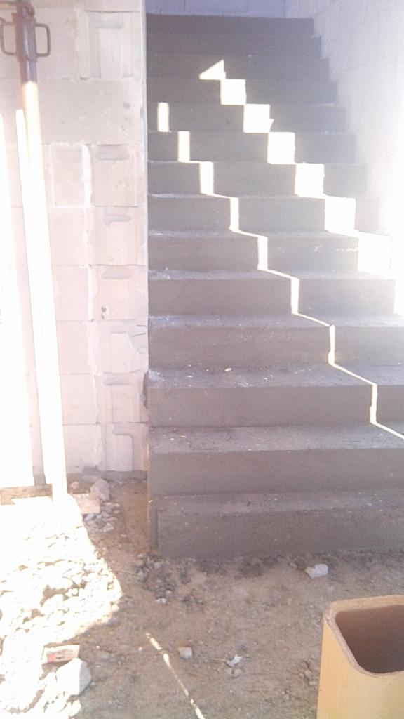 Surowe betonowe schody w trakcie budowy, z widocznymi cieniami i bloczkami betonowymi po lewej stronie, otoczone gruzem i ziemią.