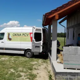 Montaż okien w surowym budynku; biały bus z otwartymi drzwiami i naklejką reklamową stoi obok budynku z zamontowanymi oknami, pracownik w niebieskiej koszulce przy oknie.