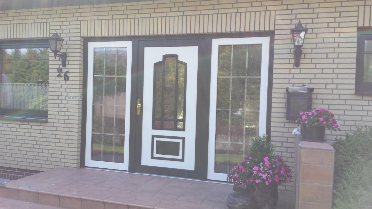 Eleganckie drzwi wejściowe z bocznymi oknami, białe obramowania, ciemny kolor drzwi, złota klamka, ceglana elewacja budynku, kwiaty w donicach przy wejściu.