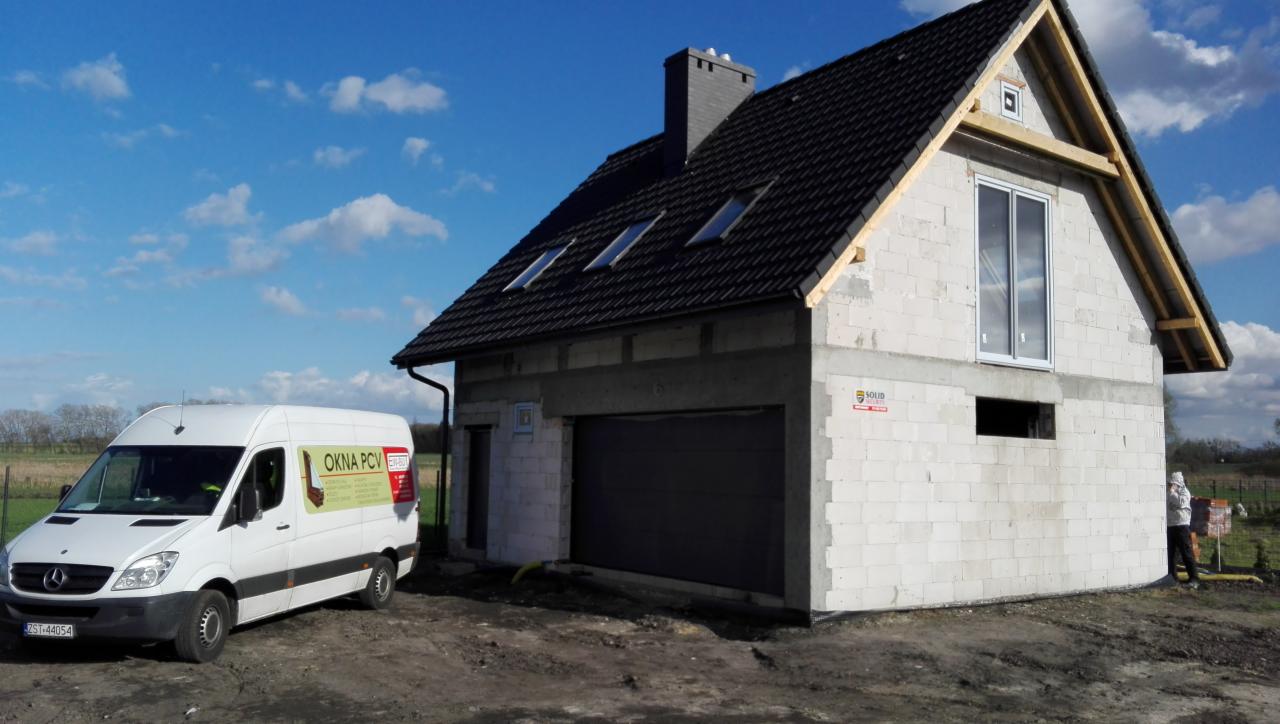 Dom w trakcie budowy z zamontowaną białą stolarką okienną, widoczny także szary segment bramy garażowej i firmowy bus z naklejką reklamową na boku.