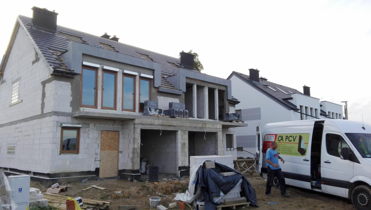 Budowa domu jednorodzinnego w trakcie montażu stolarki okiennej, widoczny biały bus z reklamą firmy, teren budowy z materiałami budowlanymi.