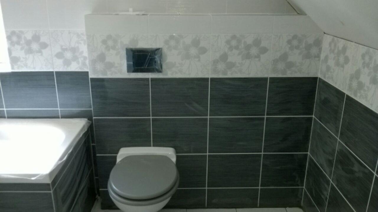 Wykończona łazienka z szarymi i białymi płytkami, wzór kwiatowy na górnej części ściany, widoczna toaleta i fragment wanny z zabezpieczeniem folią.