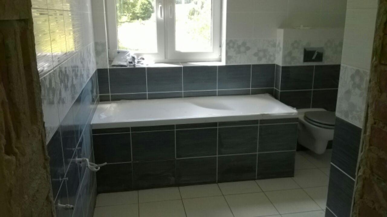 Nowoczesna łazienka z wanną obudowaną ciemnymi płytkami, częściowo wykończona, widoczne fragmenty surowej ściany i toaleta.