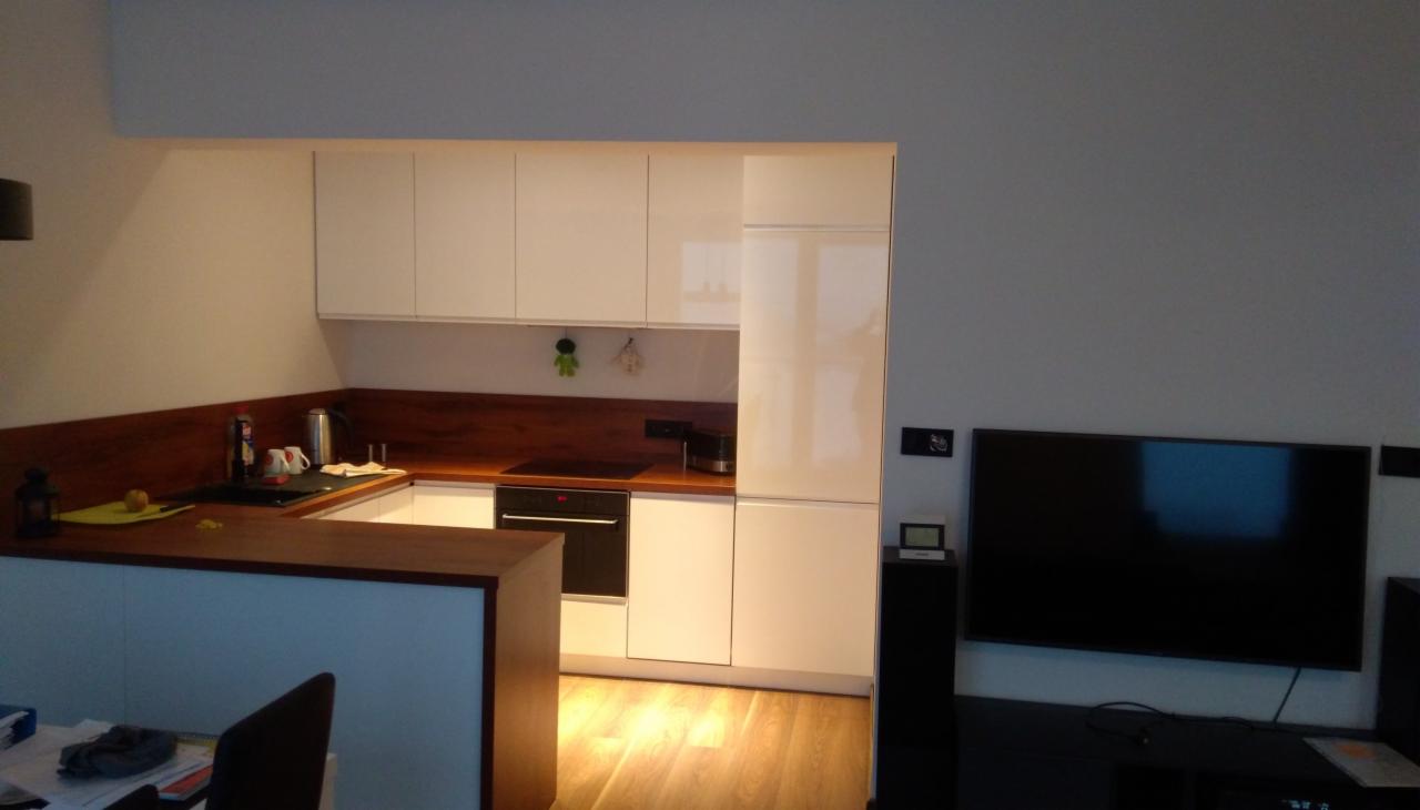 Nowoczesna kuchnia z białymi, lakierowanymi szafkami i drewnianym blatem, widok z salonu, na blacie czajnik, ekspres do kawy i akcesoria kuchenne, a w tle telewizor