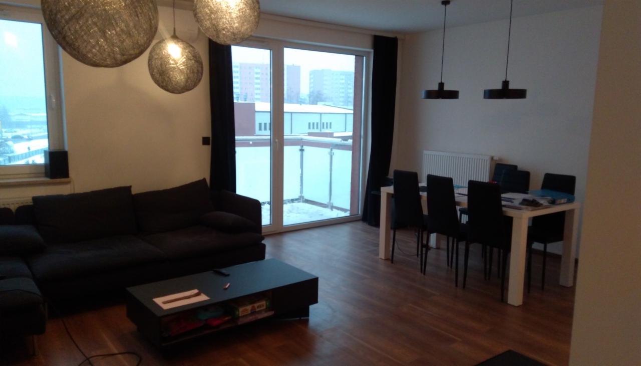 Salon z ciemną sofą narożną, czarnym stolikiem kawowym, białym stołem jadalnym z czarnymi krzesłami i trzema lampami wiszącymi w kształcie kuli, widok na balkon i budynki za oknem.