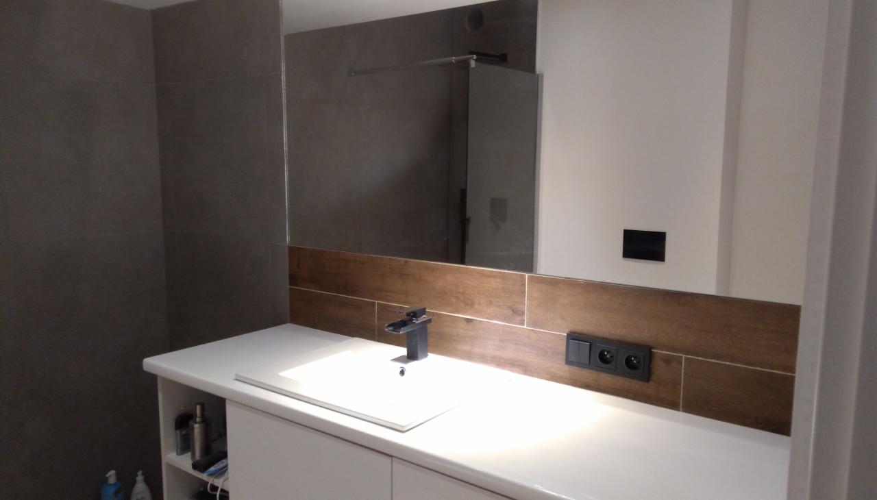 Nowoczesna łazienka z szarymi płytkami, drewnopodobnym pasem płytek nad umywalką z czarną armaturą i dużym lustrem odbijającym kabinę prysznicową.