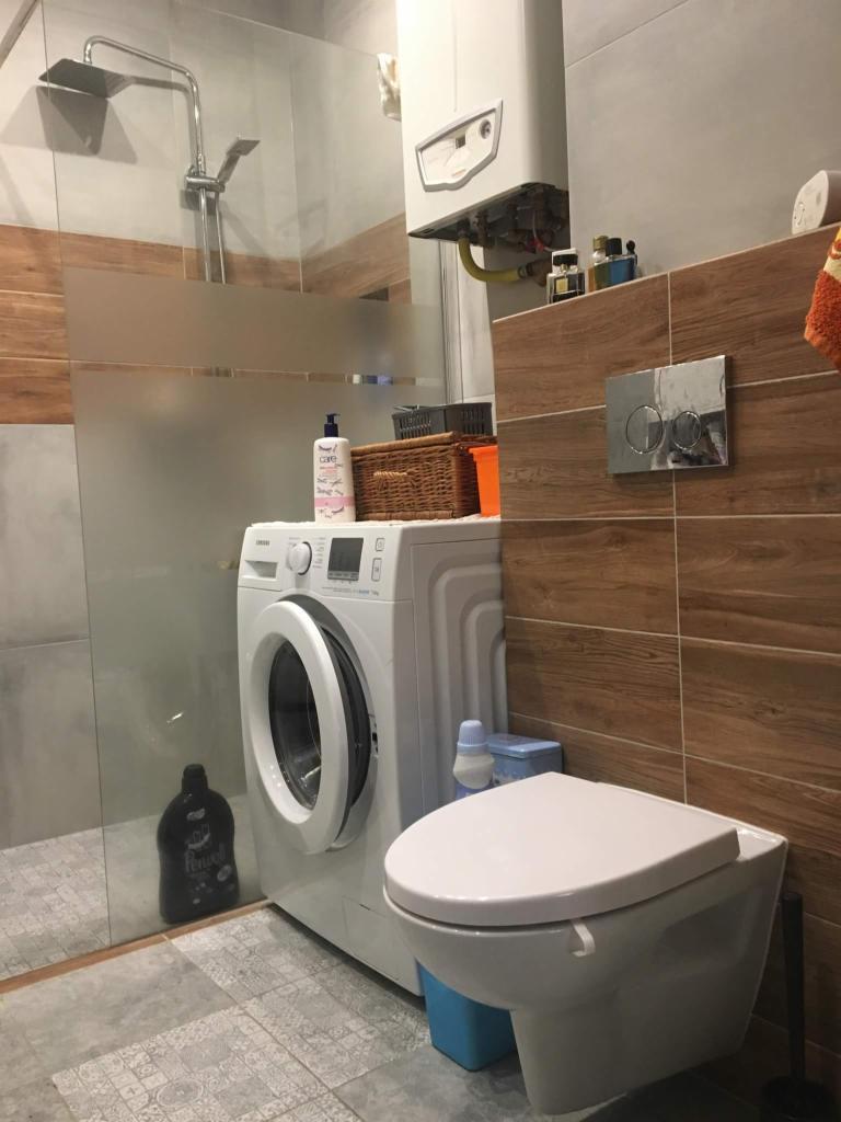 Kompaktowa łazienka z pralką obok toalety podwieszanej, brązowe płytki imitujące drewno na ścianach, szklana kabina prysznicowa, wiszący piec gazowy.