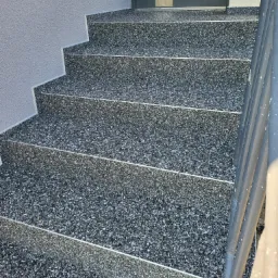 Schody zewnętrzne pokryte szarą, drobnoziarnistą posadzką żywiczną z aluminiowymi listwami krawędziowymi, widoczne z góry pod kątem, z fragmentem szarej ściany i metalowej barierki.