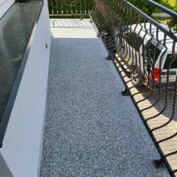 Szary taras z żywicy epoksydowej na balkonie z ozdobną metalową balustradą, widok z góry, z fragmentem zaparkowanego samochodu w tle.