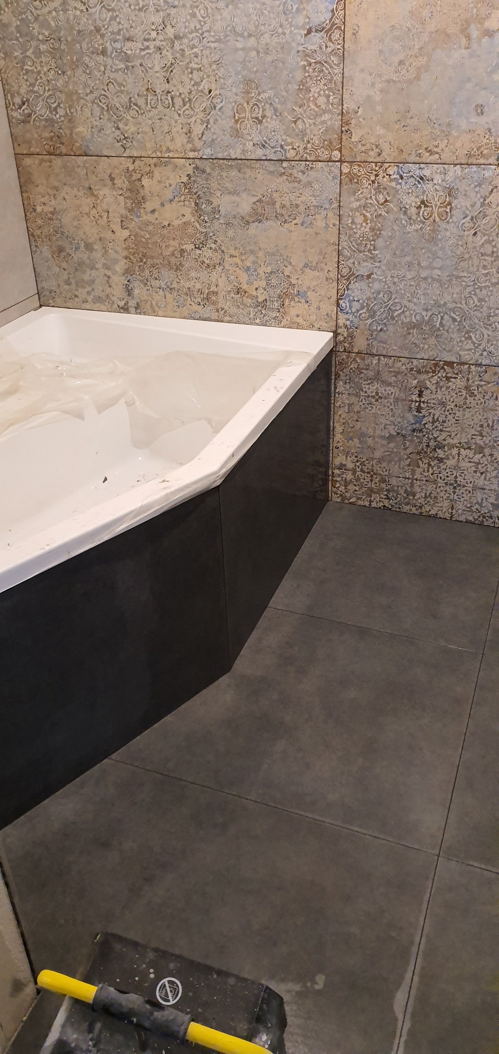 Wykończona łazienka z wanną obudowaną czarnymi płytami i ozdobnymi płytkami ściennymi w stylu vintage, widoczna szara podłoga z płytek i fragment narzędzia glazurniczego w rogu.