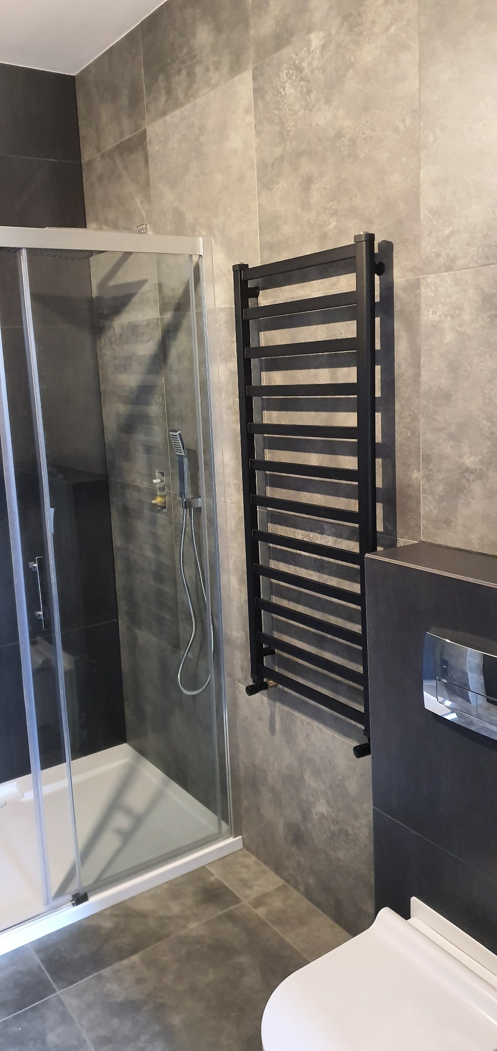 Szara łazienka z kabiną prysznicową z przesuwnymi drzwiami, czarnym grzejnikiem drabinkowym i podwieszaną toaletą.