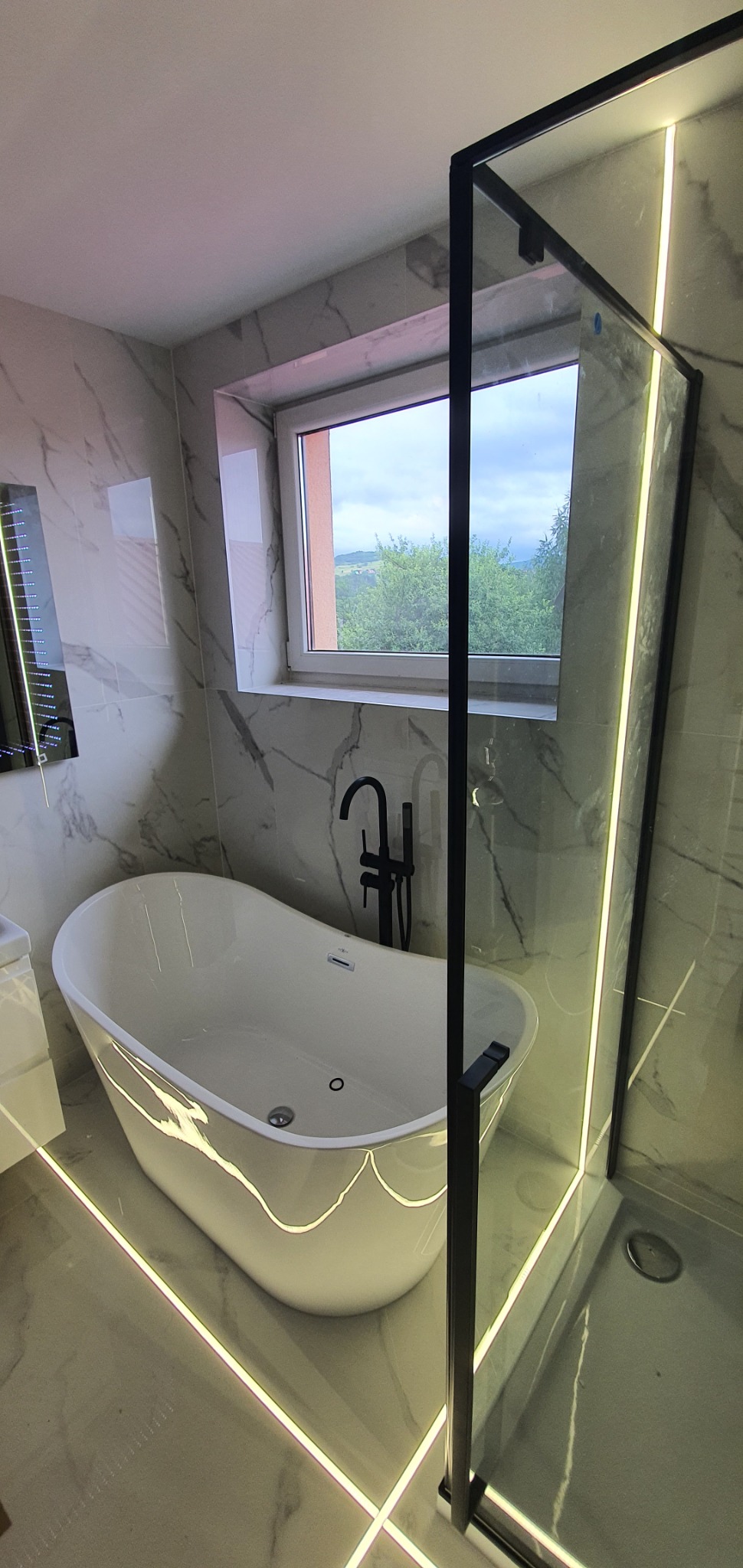 Nowoczesna łazienka z wolnostojącą wanną, czarną armaturą i kabiną prysznicową ze szklaną ścianą, podświetlona listwami LED w podłodze i przy kabinie, z widokiem na zielone wzgórza za oknem.