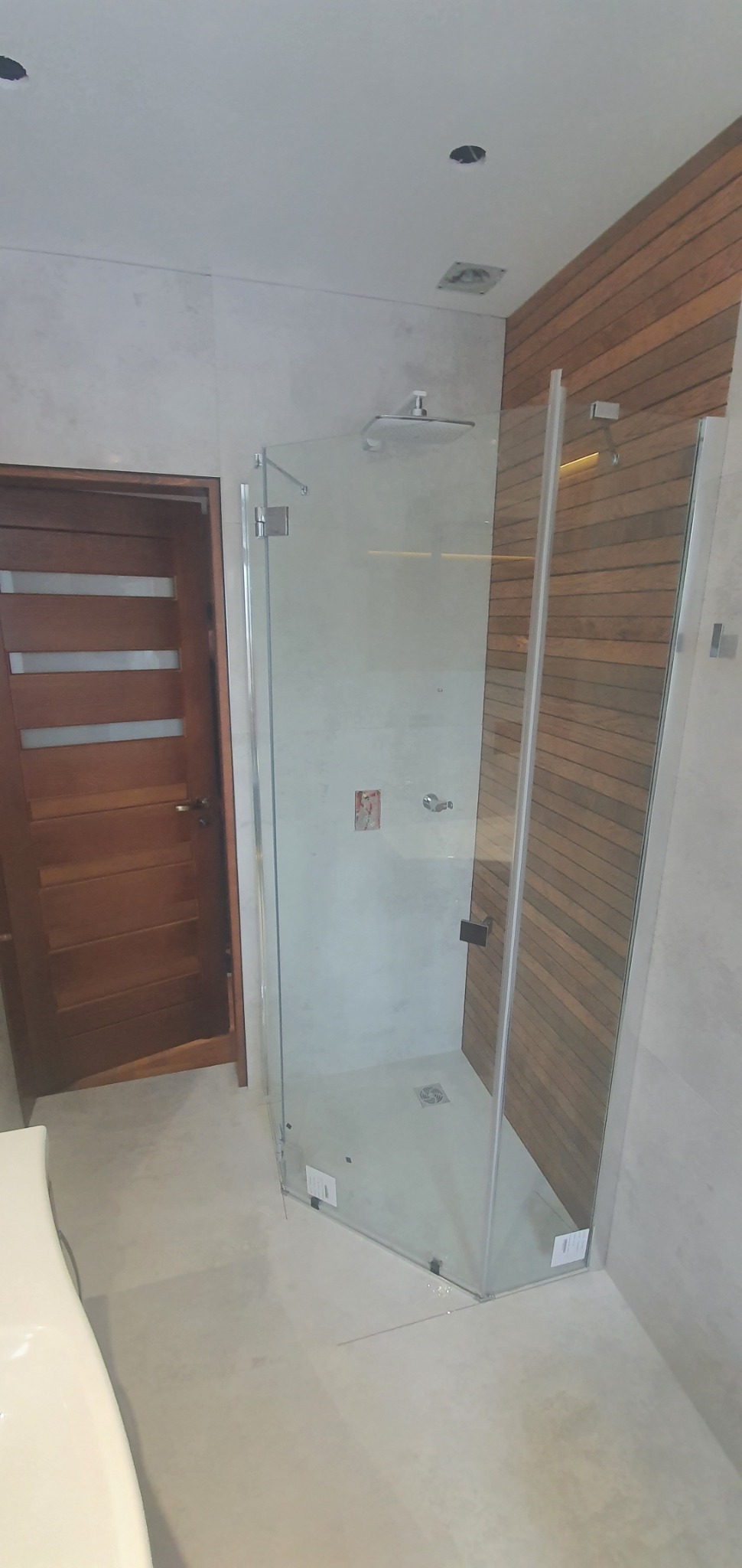 Nowoczesna łazienka z kabiną prysznicową typu walk-in z drewnianą ścianą, widoczne niedokończone otwory w suficie na oświetlenie.