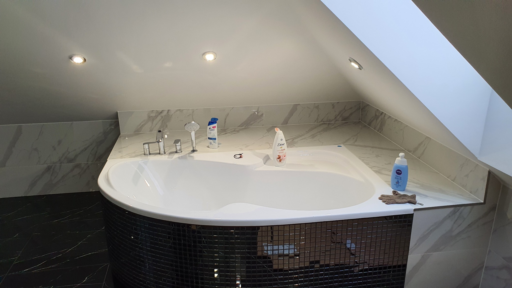 Wanna w łazience na poddaszu, obudowana mozaiką, z armaturą i kosmetykami. Ściany wykończone płytkami imitującymi marmur.