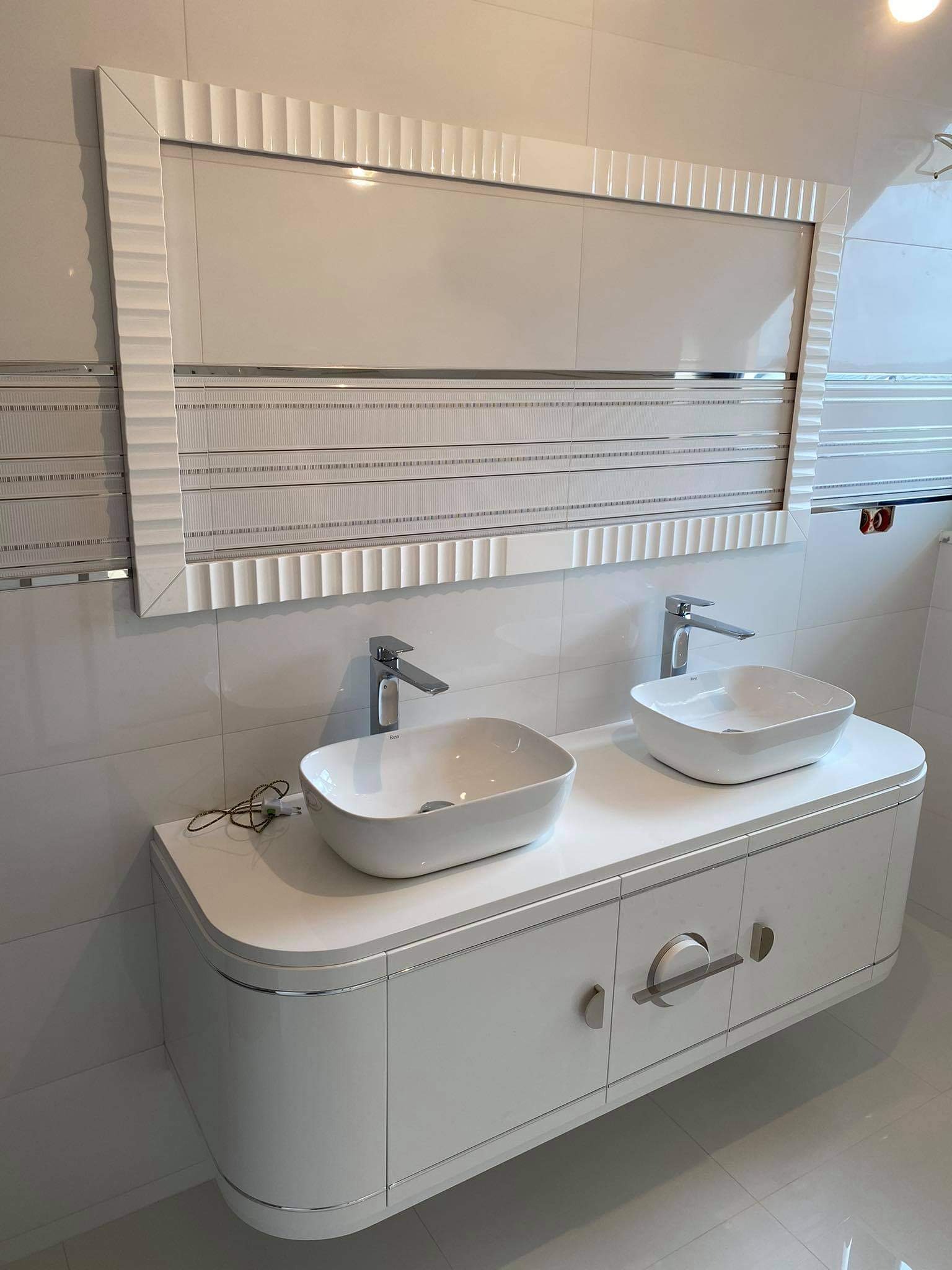 Nowoczesna łazienka z podwójną umywalką nablatową na białej szafce z zaokrąglonymi krawędziami i lustrem w dekoracyjnej ramie, białe płytki na ścianach i podłodze.