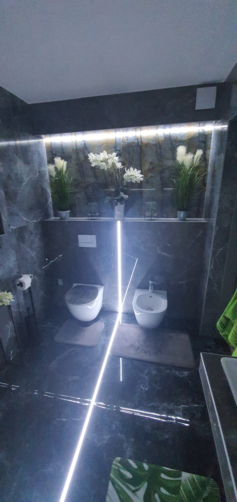 Nowoczesna łazienka wykończona marmurem z podświetleniem LED, z wiszącą toaletą i bidetem na tle dekoracyjnych kwiatów w doniczkach.