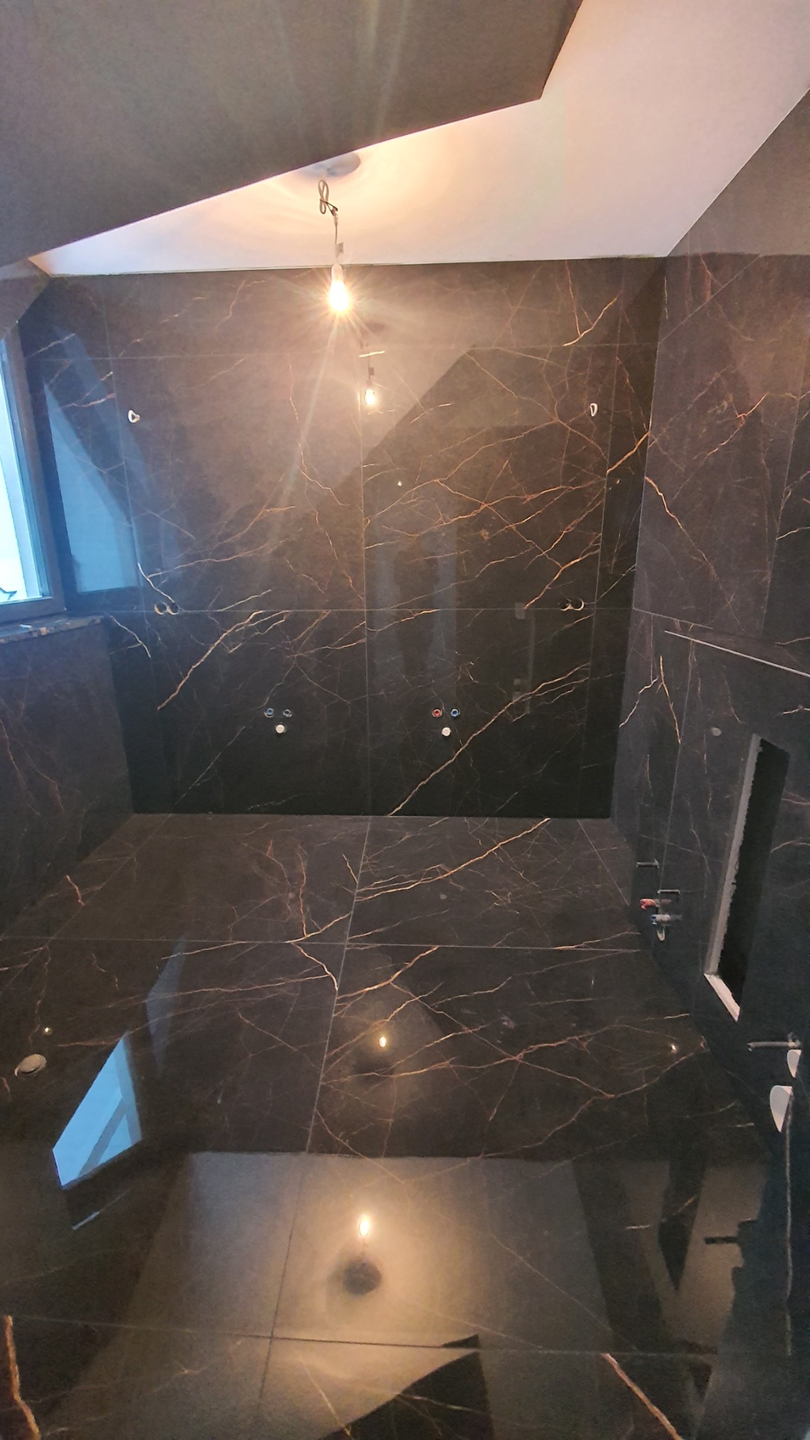 Wnętrze łazienki w trakcie wykańczania, ściany i podłoga wyłożone czarnymi płytkami z wyraźnym złotym żyłkowaniem, widoczne otwory na armaturę i instalacje, sufit częściowo podwieszany, oświetlenie...