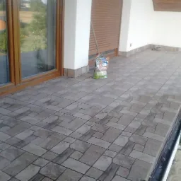 Wyłożony płytkami taras z widocznym opakowaniem fugi, z brązowymi ramami okiennymi i roletami, częściowo widoczną drabiną i zielenią w tle.