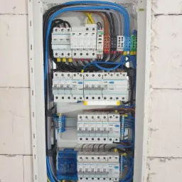 Rozdzielnica elektryczna z zabezpieczeniami i okablowaniem, zamontowana w nowym budynku w Głogowie, widoczna na tle ściany z bloczków. Widoczne bezpieczniki firmy Hager i IDEAL.