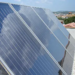 Kolektory słoneczne na dachu pokrytym żwirem, odbijające błękitne niebo z chmurami, z widokiem na zabudowania w oddali.