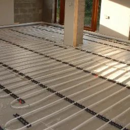 Białe rurki ogrzewania podłogowego zamocowane w czarnych uchwytach, zalane w wylewce betonowej, widok z góry w surowym stanie.