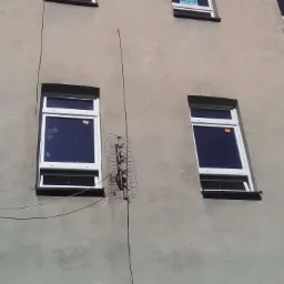 Fasada budynku z kilkoma białymi oknami, na jednym z okien naklejka informacyjna, pomiędzy oknami zamontowana antena telewizyjna.