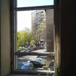 Montaż okna. Widok z wnętrza mieszkania na okno z widocznym blokiem mieszkalnym i zaparkowanymi samochodami. Na parapecie leżą narzędzia: poziomica i młotek.