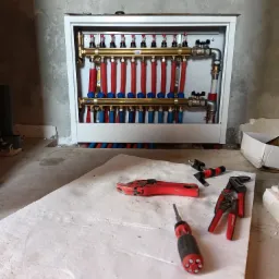 Rozdzielacz ogrzewania podłogowego w białej obudowie, widoczne czerwone i niebieskie rury, na pierwszym planie narzędzia: obcinak do rur, zaciskarka i śrubokręt na białym styropianie.