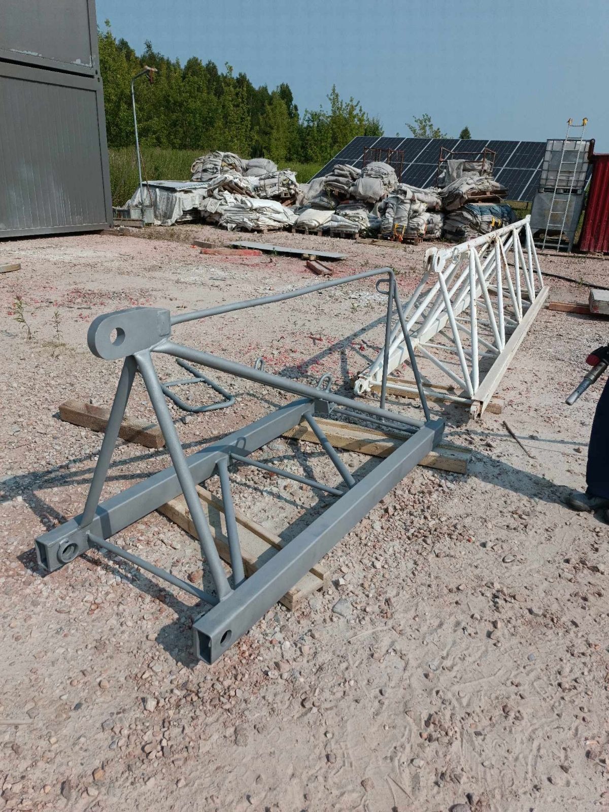 Dwie metalowe konstrukcje kratowe, jedna szara, druga biała, leżące na ziemi z gruzem i drewnianymi podkładkami, w tle panele słoneczne i worki z materiałem.