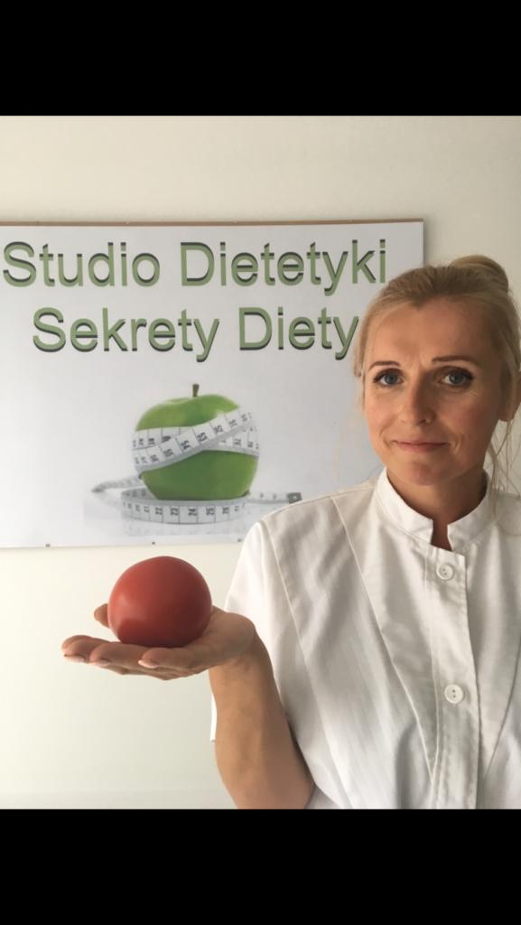Dietetyczka w białym uniformie trzymająca pomidora na dłoni, w tle tablica z napisem Studio Dietetyki Sekrety Diety i jabłko owinięte miarką.