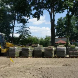 Żółta koparko-ładowarka obok palet z rolkami trawy na przygotowanym podłożu, w tle drzewa i budynki.