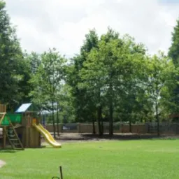 Drewniany plac zabaw z wieżą, zjeżdżalnią i ścianką wspinaczkową, otoczony zielonym trawnikiem i drzewami, ogrodzony niskim kamiennym murkiem.