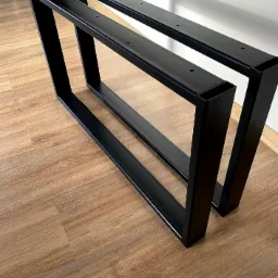Dwie czarne, metalowe nogi stołowe w kształcie trapezu, wykonane z profili zamkniętych, leżące na podłodze imitującej drewno.