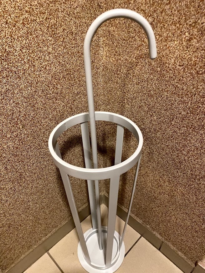 Biały, metalowy stojak na parasole w kształcie otwartego kosza z uchwytem przypominającym laskę, umieszczony w rogu pomieszczenia z beżową, teksturowaną ścianą.