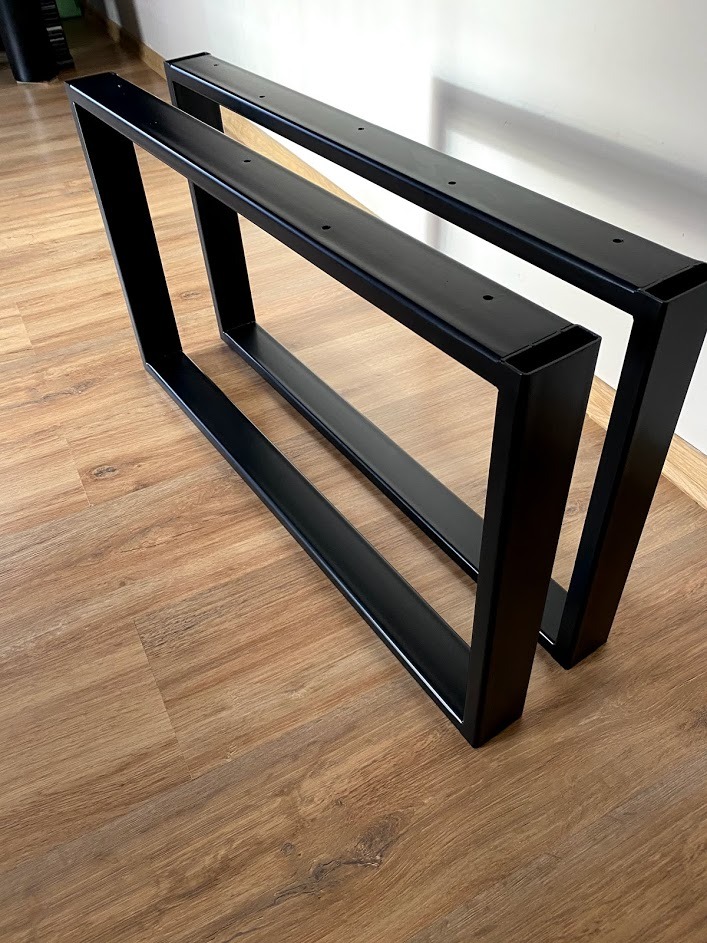 Dwie czarne, metalowe nogi stołowe w kształcie trapezu, wykonane z profili zamkniętych, leżące na podłodze imitującej drewno.