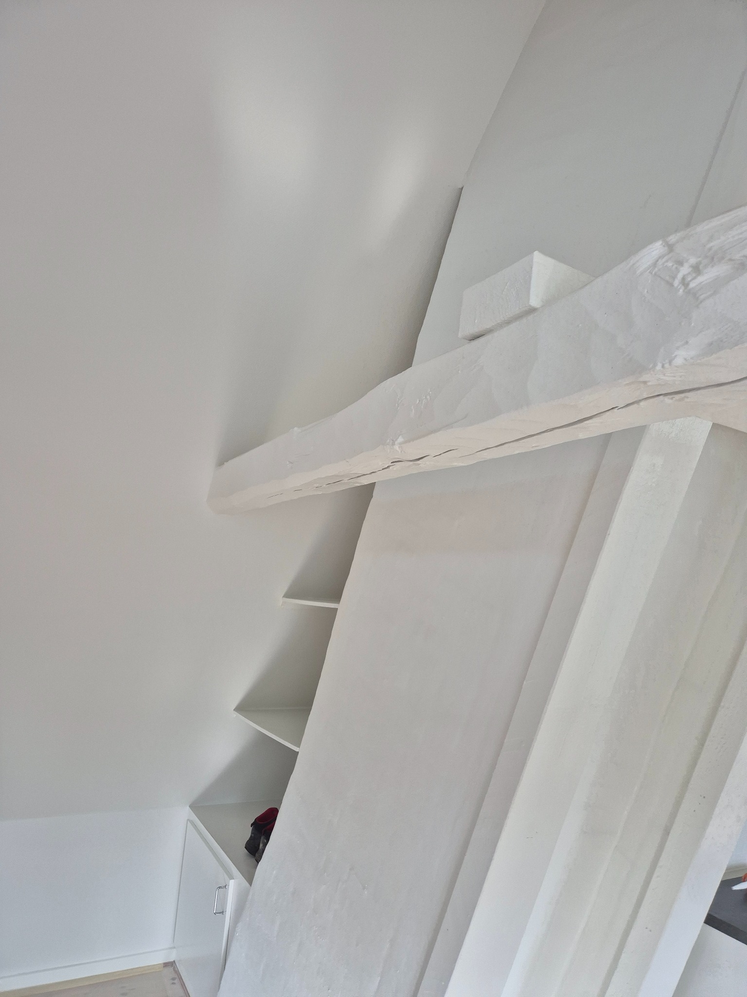 Białe wnętrze poddasza ze skośnym sufitem, drewnianą belką i wbudowanymi półkami. Minimalistyczny design, jasne oświetlenie, widok z dołu.