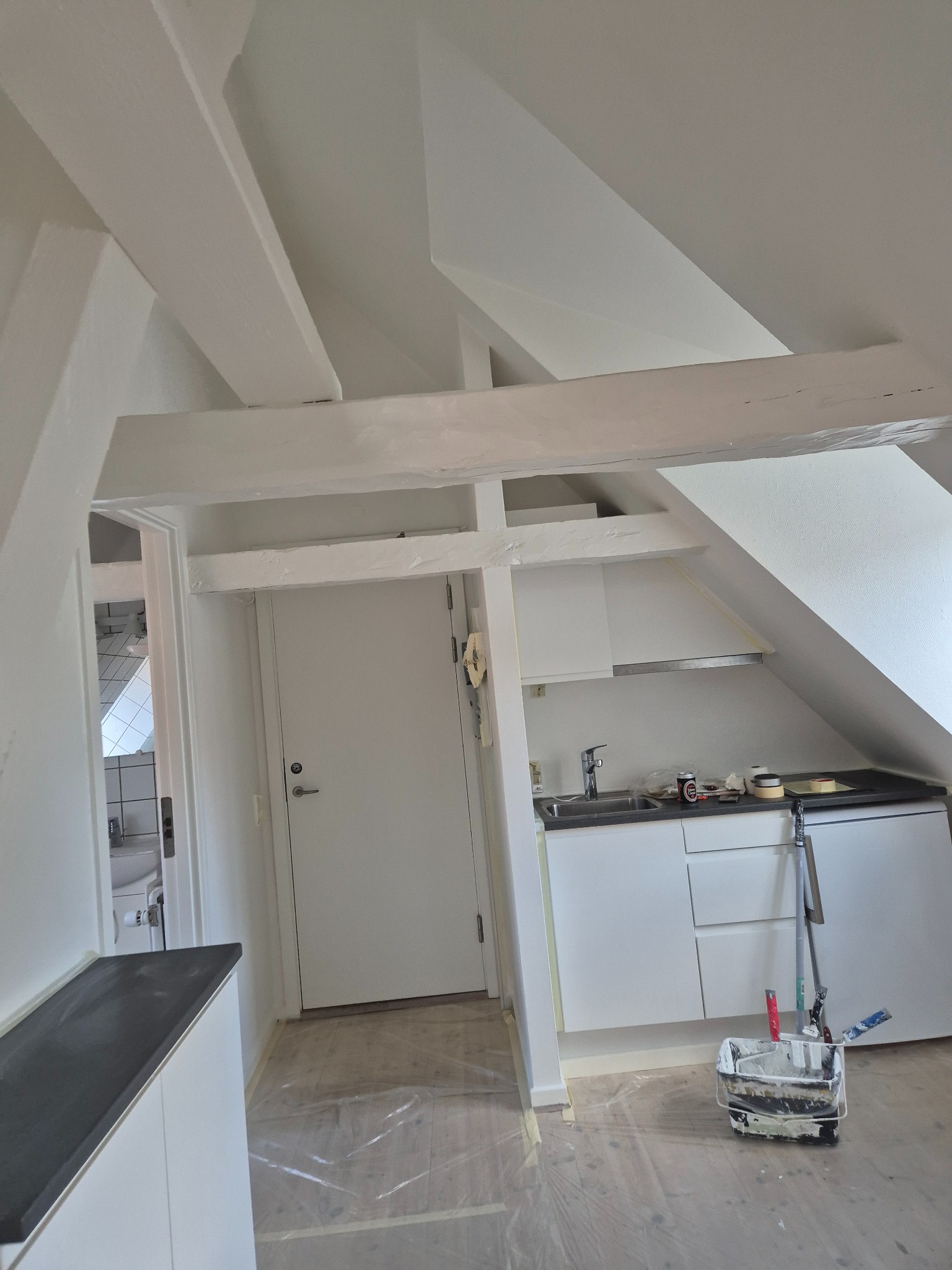 Białe wnętrze poddasza z drewnianymi belkami po odświeżeniu, widoczna kuchnia i drzwi, podłoga zabezpieczona folią, narzędzia malarskie w wiadrze.