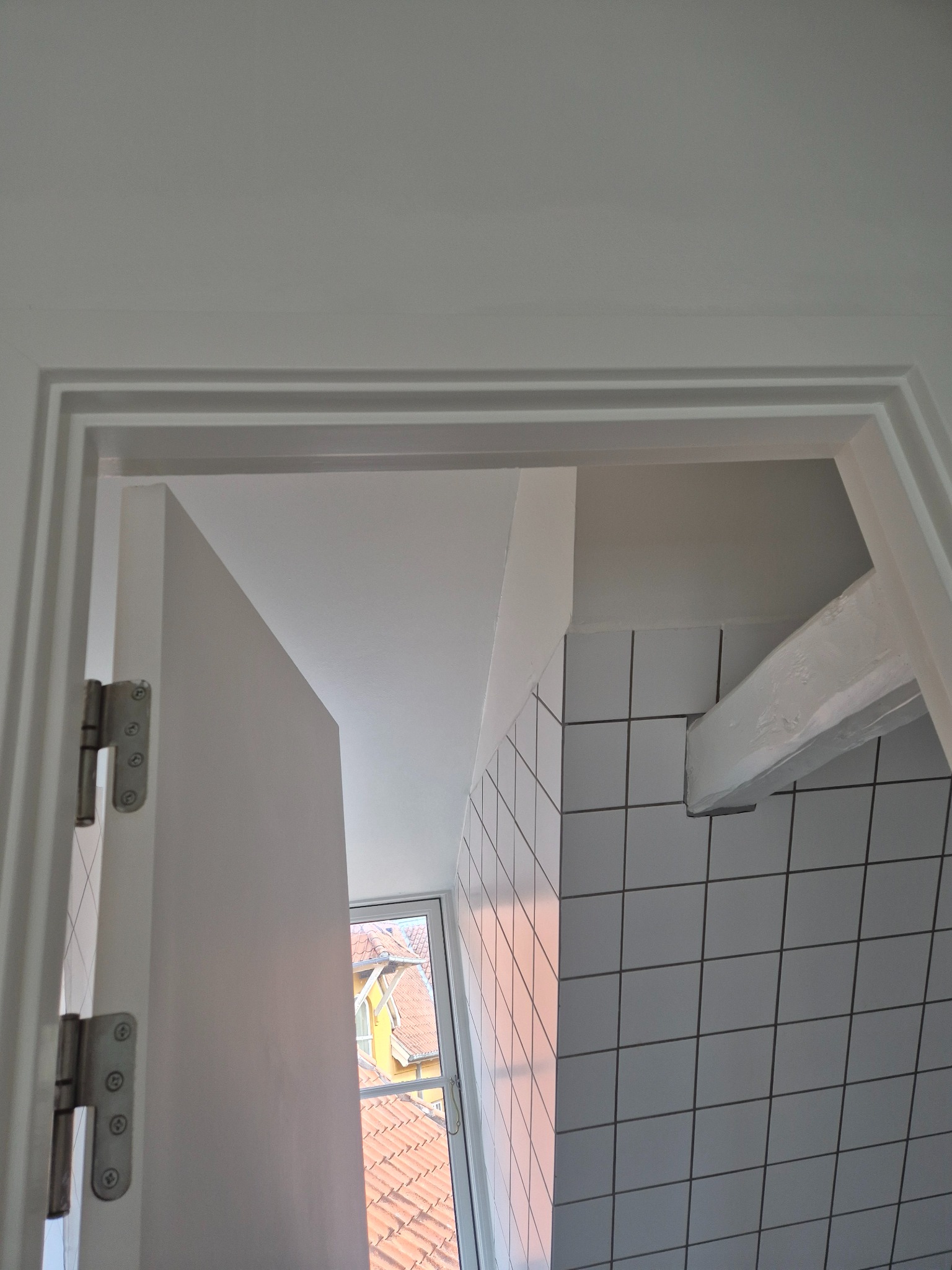 Biała, częściowo otwarta łazienka z kwadratowymi płytkami, widok zza drzwi. Widoczny fragment okna i dachu budynku na zewnątrz. Minimalistyczny design.