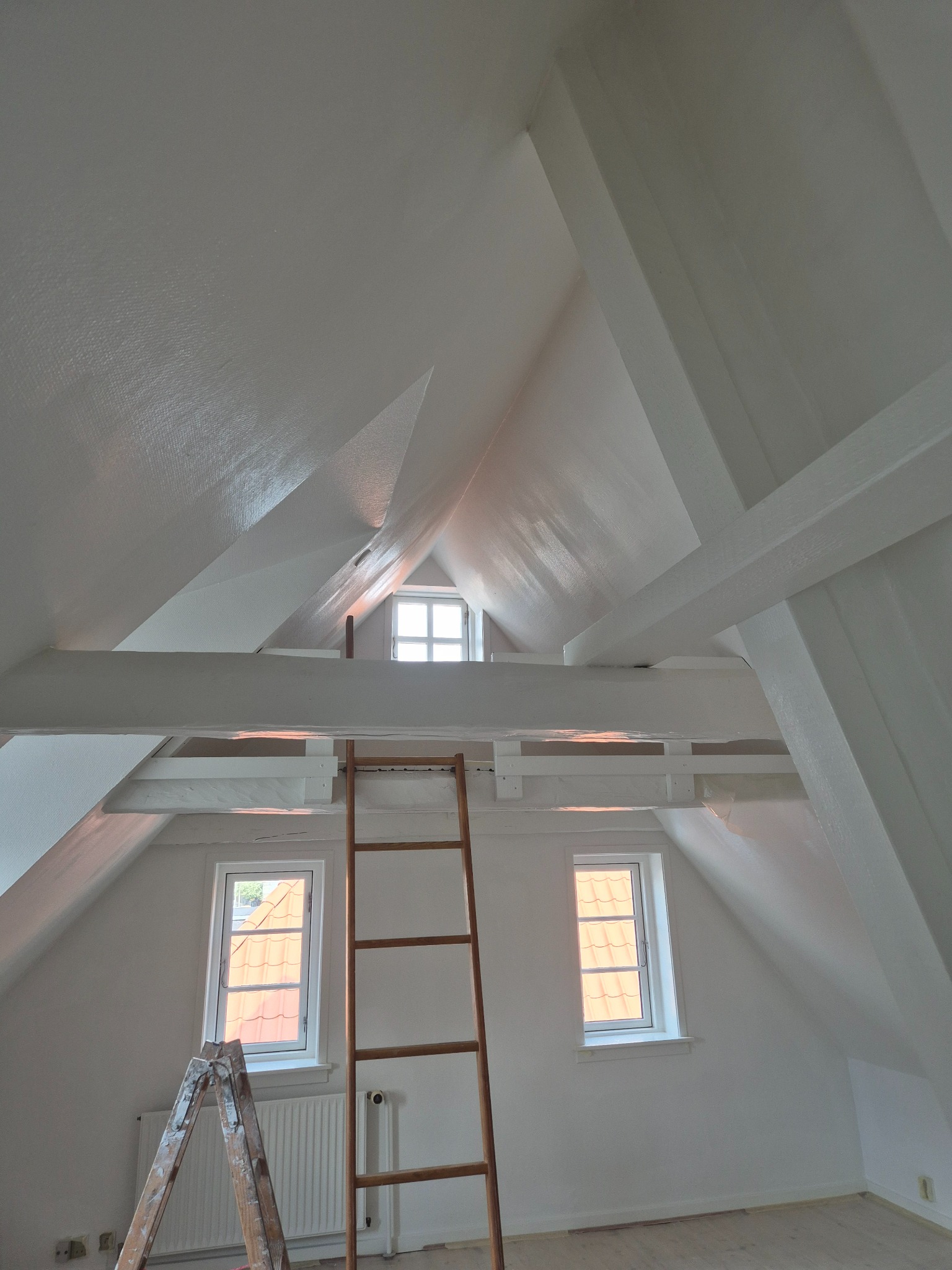 Białe poddasze po remoncie z drewnianą drabiną, widoczne okna i skosy. Świeżo pomalowane ściany i sufit. Ujęcie z dołu, podkreślające geometrię pomieszczenia.