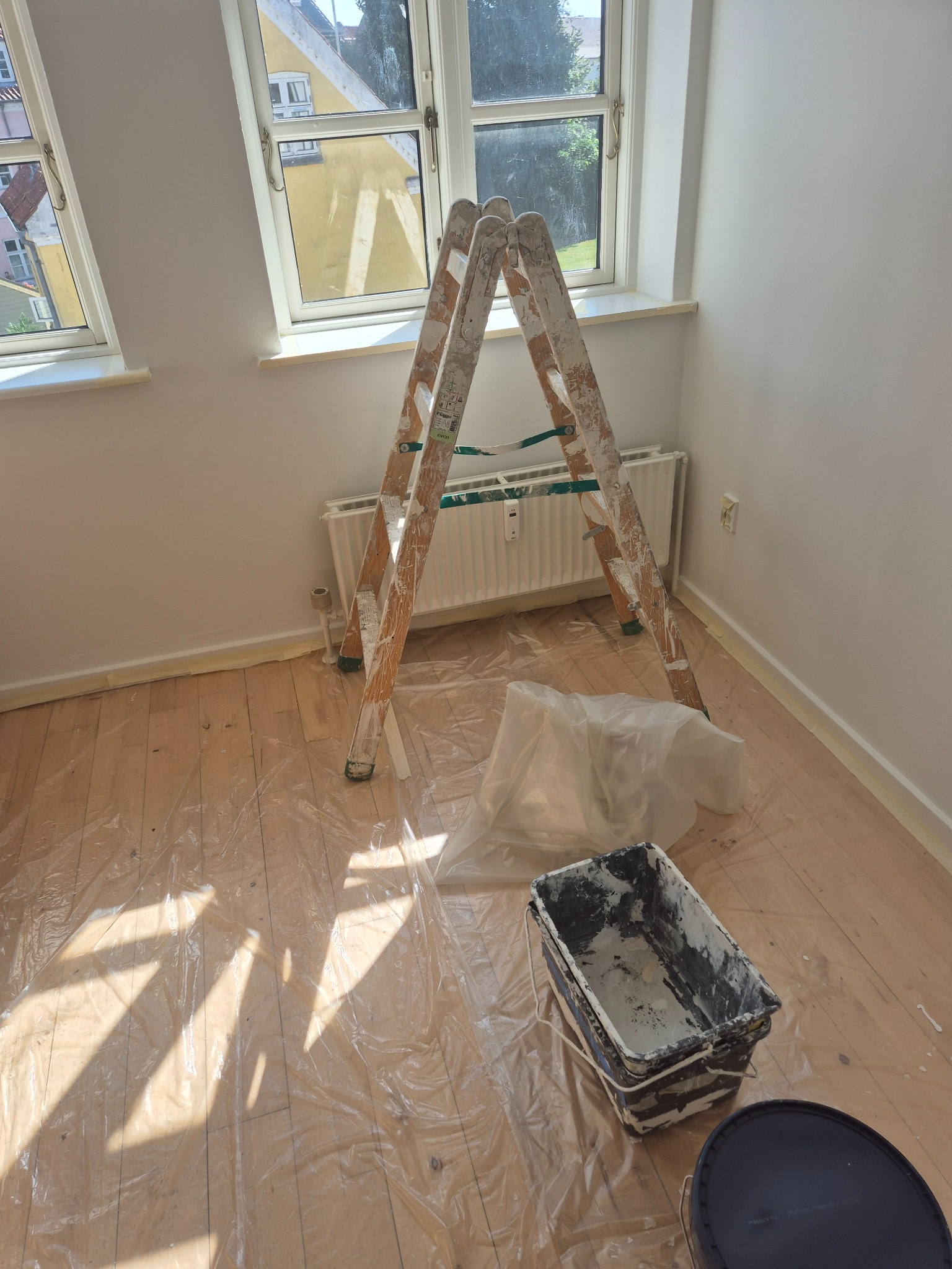Pokój po odświeżeniu ścian na biało. Widoczna drabina malarska, wiadro z farbą i folia ochronna na podłodze. Okna w tle wpuszczają naturalne światło.