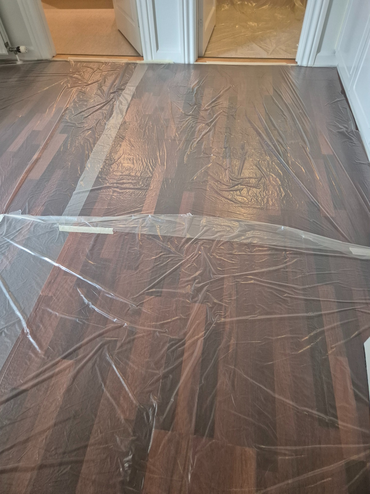 Parkiet zabezpieczony folią malarską przed pracami remontowymi. Widoczne białe listwy przypodłogowe i fragment otwartych drzwi do innego pomieszczenia.