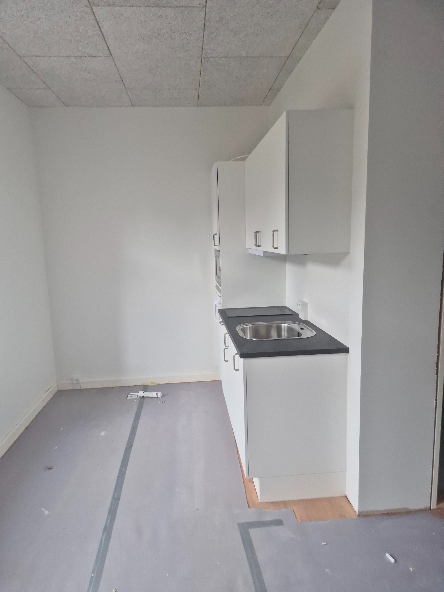 Nowoczesna, minimalistyczna kuchnia z białymi szafkami i szarym blatem, surowy stan wykończenia podłogi, widok na sufit z płyt.