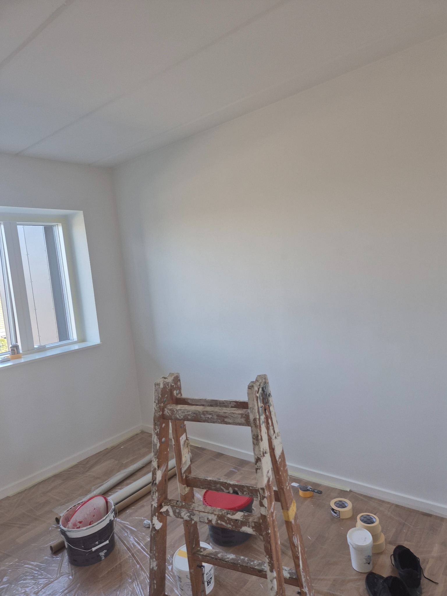 Białe ściany po malowaniu w pokoju z oknem. Drewniana drabina malarska, wiadro z farbą i taśmy malarskie na podłodze zabezpieczonej folią. Widok z dołu.