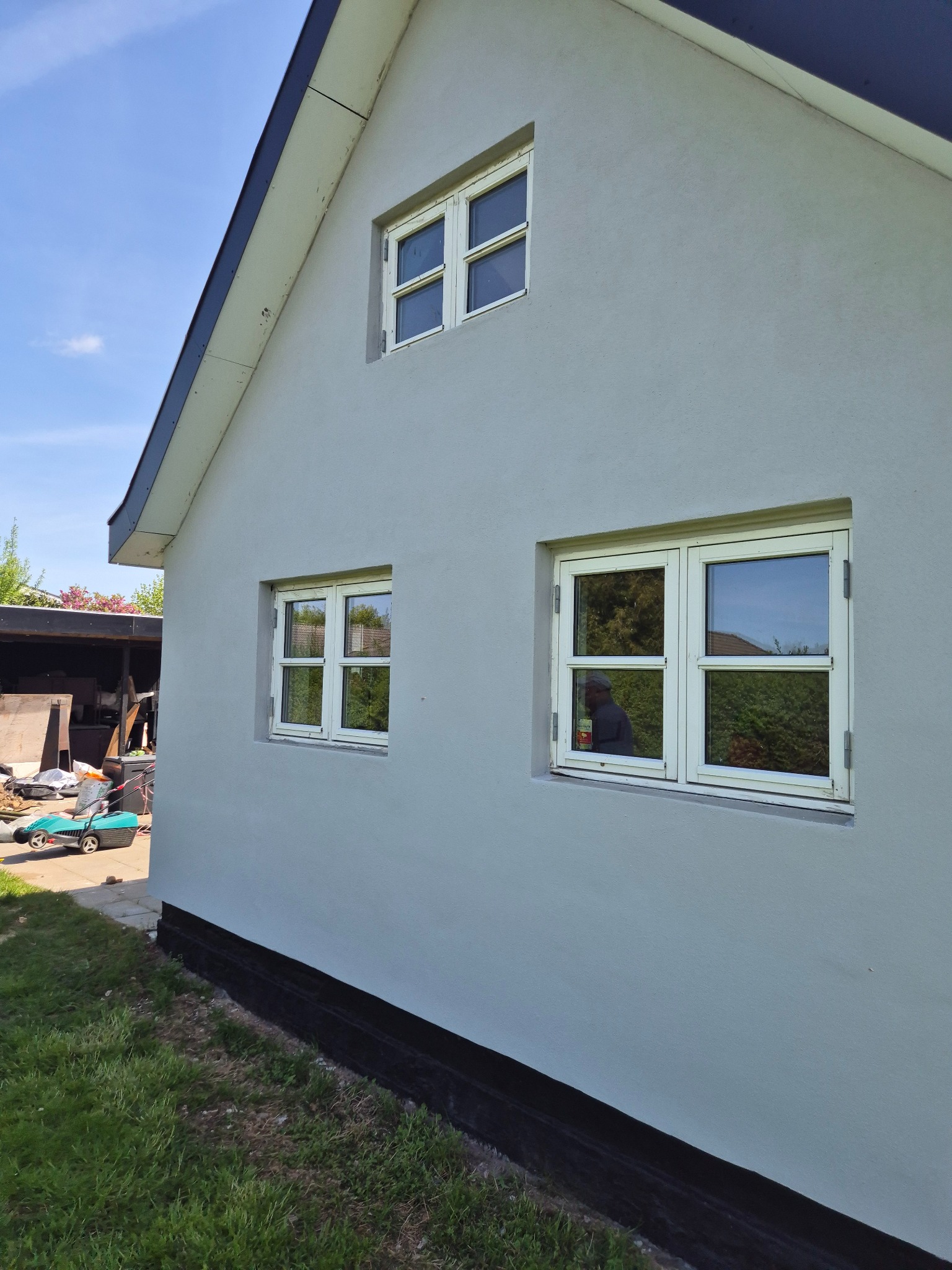 Świeżo pomalowana elewacja domu z białymi oknami i czarną podmurówką, widok z boku na tle zielonego trawnika i błękitnego nieba. W oknach odbija się osoba.